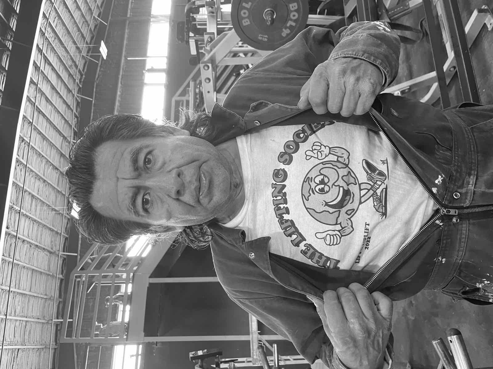 A man in a gym pulling open his jacket to reveal a white t-shirt printed with The Lifting Society logo — a smiling globe mascot giving a thumbs up and flexing, above the tagline 'LIFT AND UPLIFT 2022'.